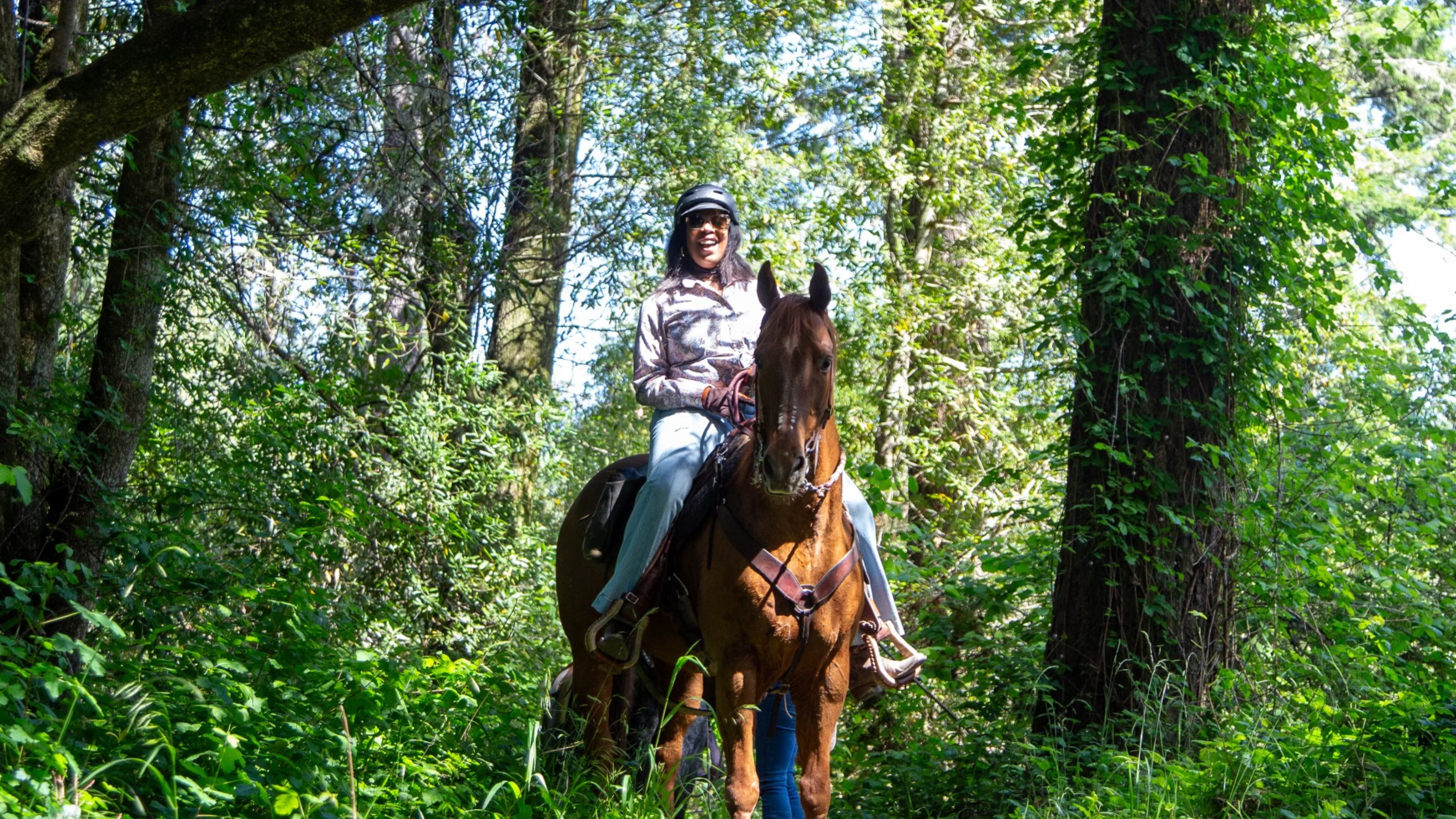 Rue Mapp smiling while riding a brown horse in the woods